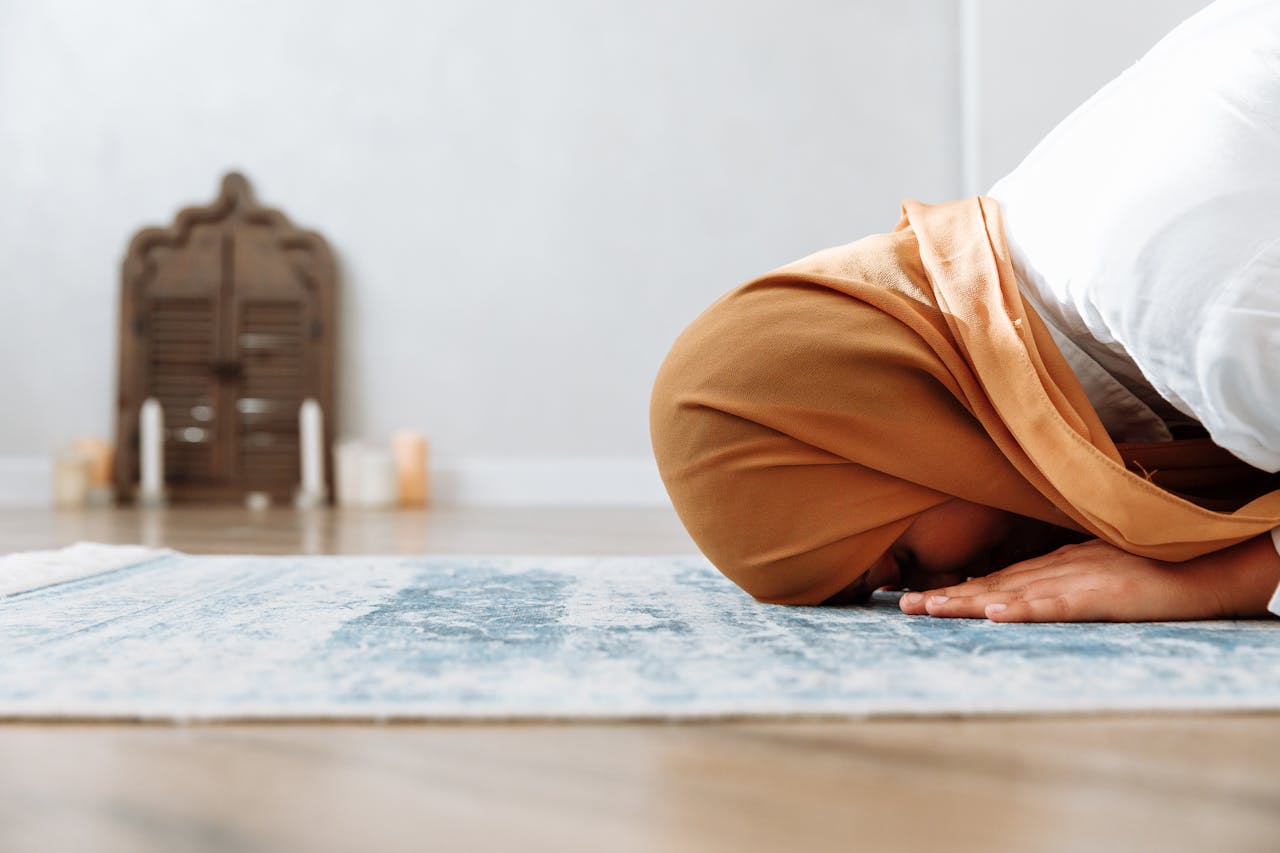 A woman in a hijab kneels in prayer on a rug indoors, with focus on devotion.